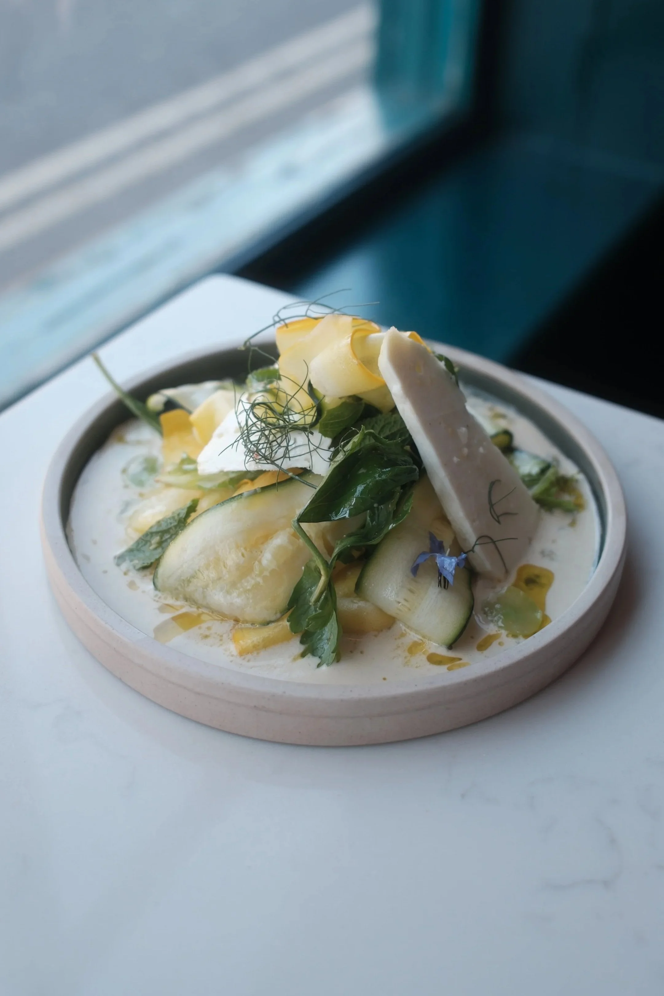 Close-up of a bowl of creamy soup garnished with vegetables, herbs, and cheese, placed on a white surface near a window.