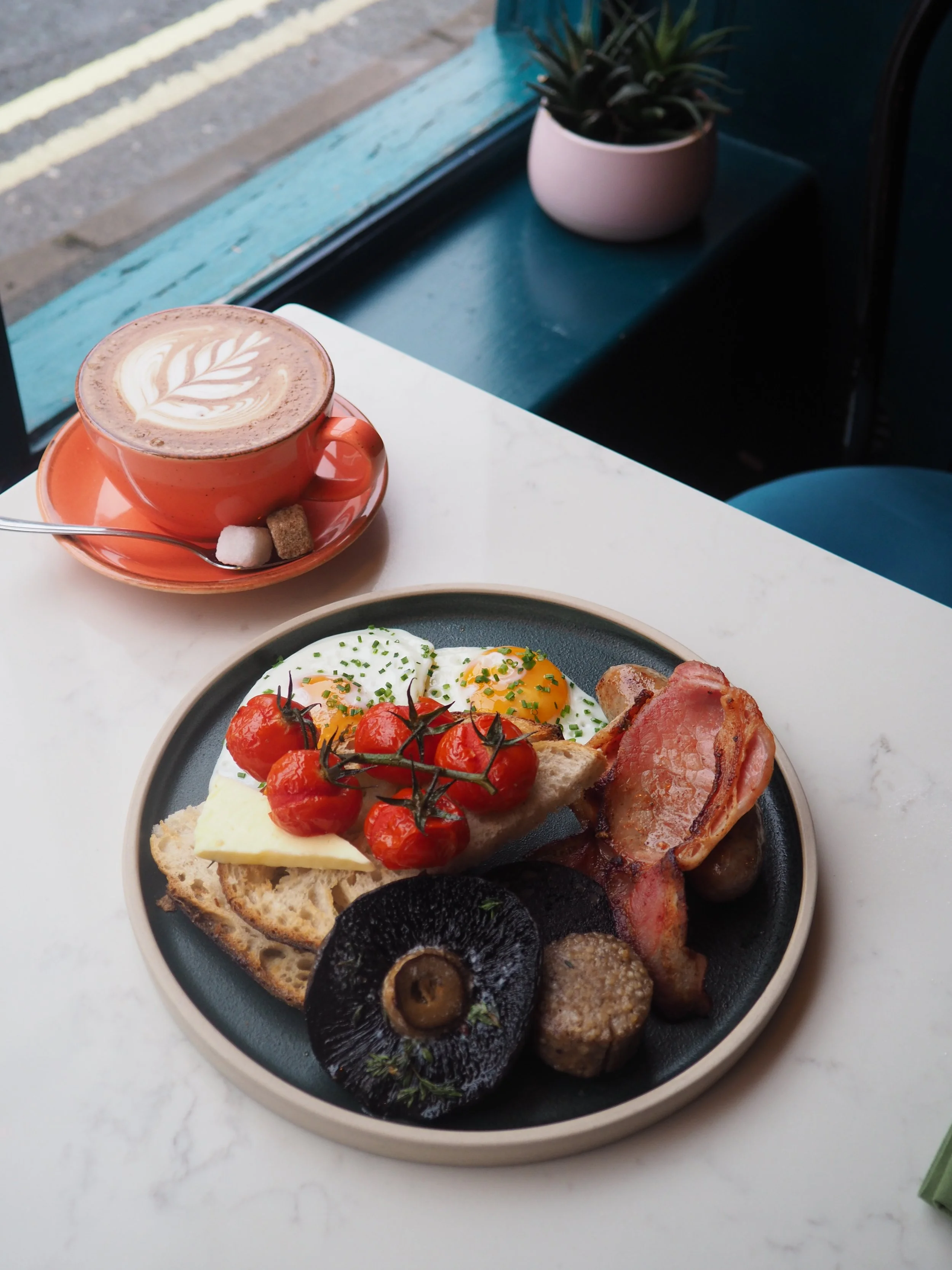 A plate of breakfast food including eggs, cherry tomatoes, bacon, sausage, toast with butter, and black pudding, with black mushrooms garnishing the plate. A cup of coffee with latte art and two sugar cubes sits on a saucer next to it, on a white table near a window with a plant in the background.