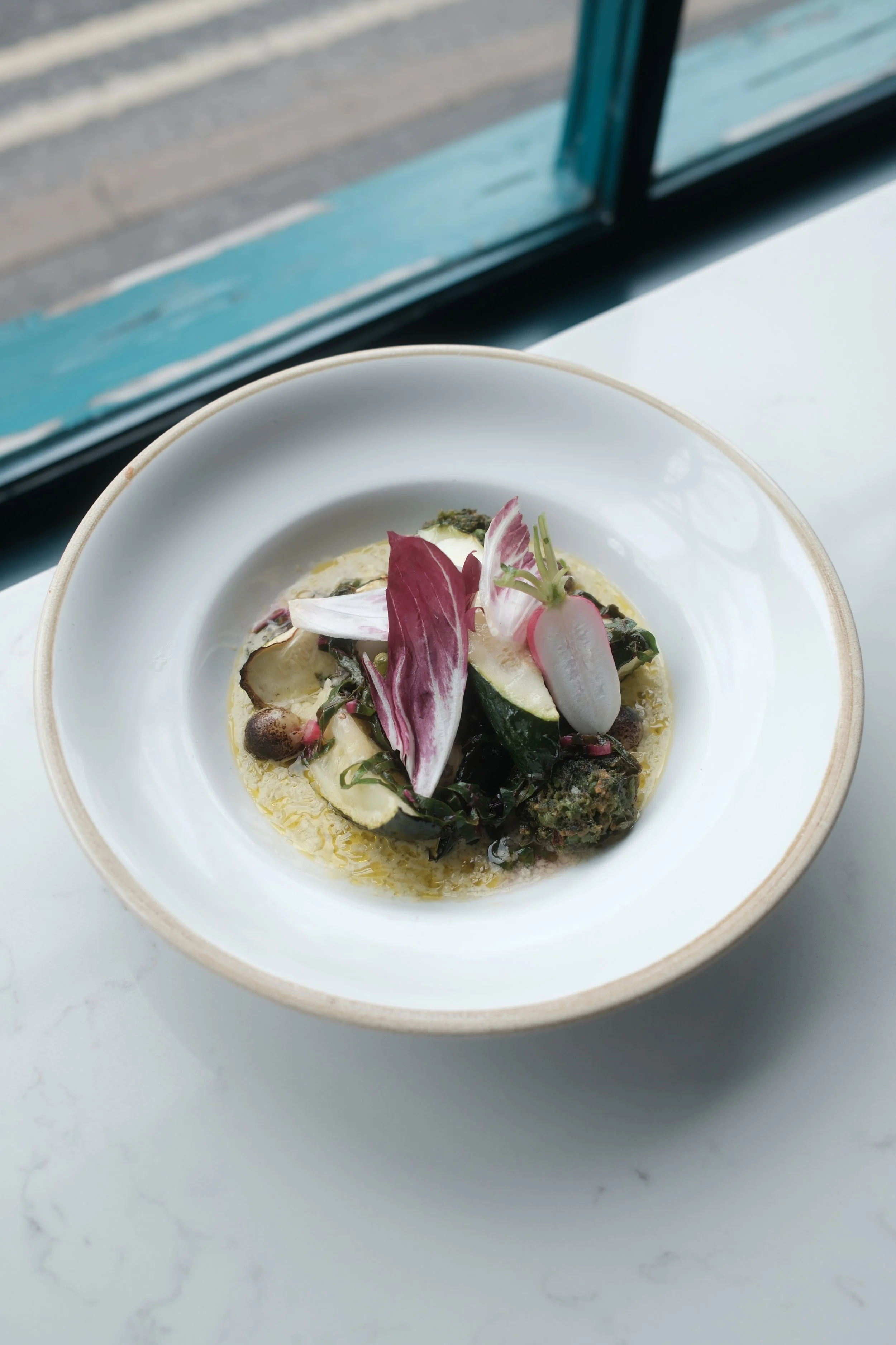 A bowl of soup garnished with radish slices, edible flowers, and herbs, placed on a white surface next to a window.