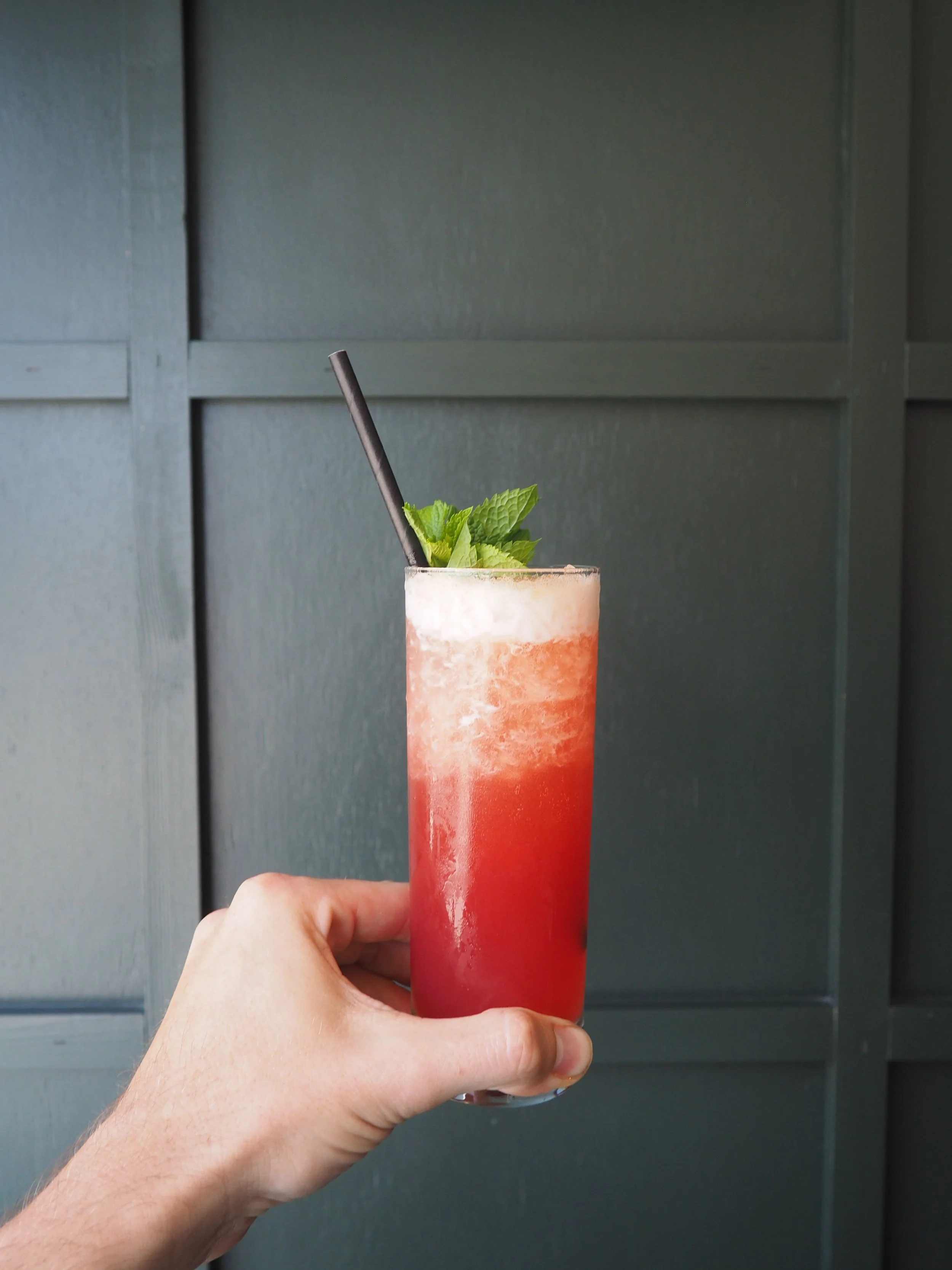 A hand holding a tall glass of frothy, layered pink and white beverage garnished with fresh mint leaves and a black straw.