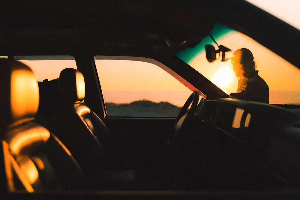 The interior of a vehicle at sunset, with a person standing outside near the front of the car.