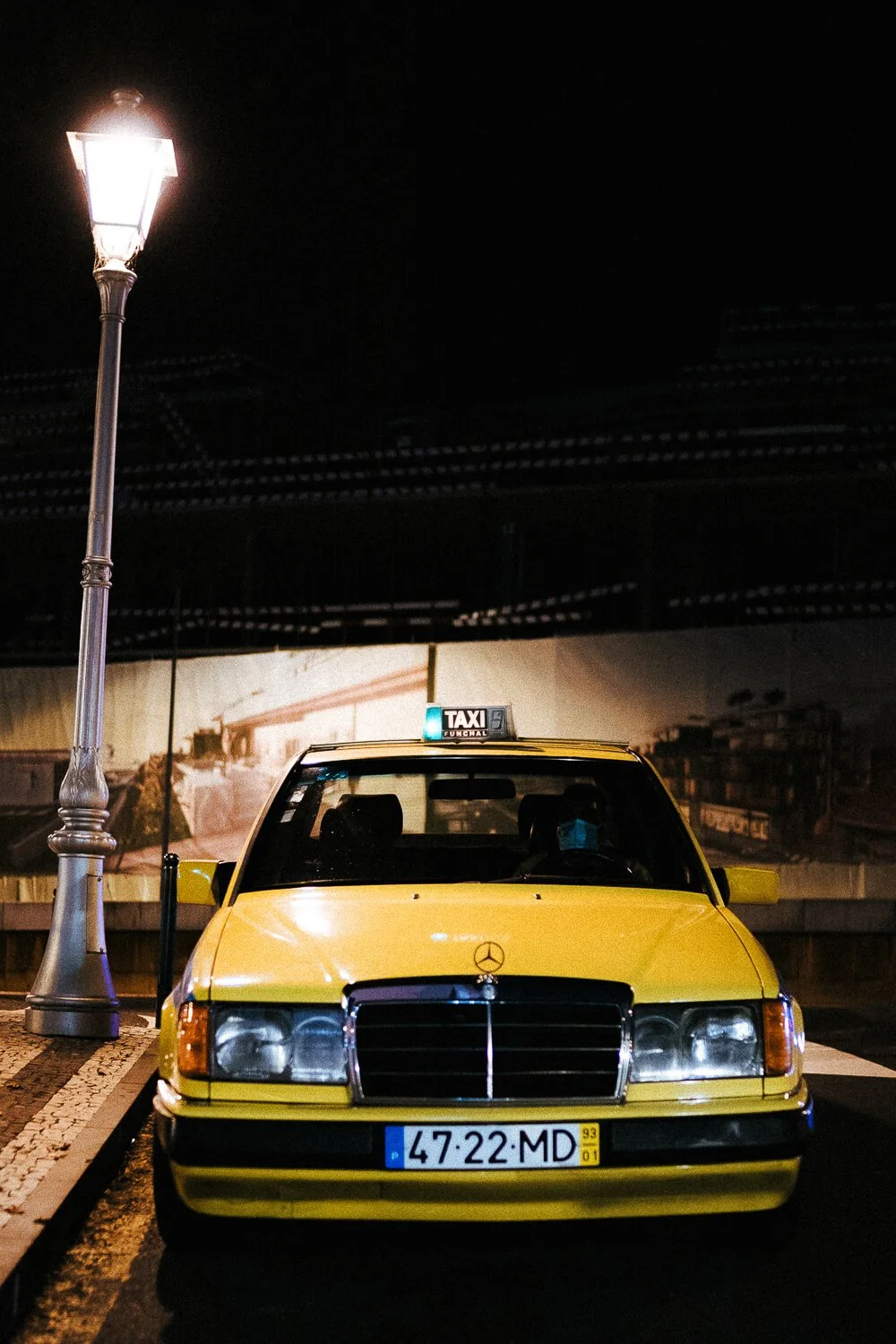 A yellow vintage Mercedes-Benz taxi parked at night near a street lamp, with a dark sky and blurred cityscape in the background.