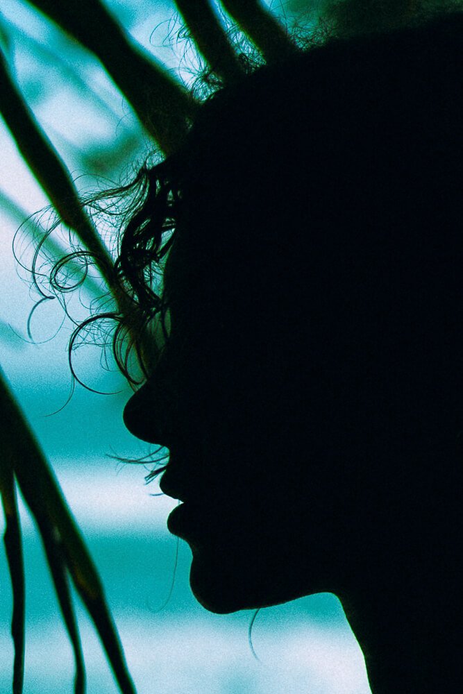 Silhouette of a person's face in profile with curly hair, against a background of greenish-blue natural light and foliage.