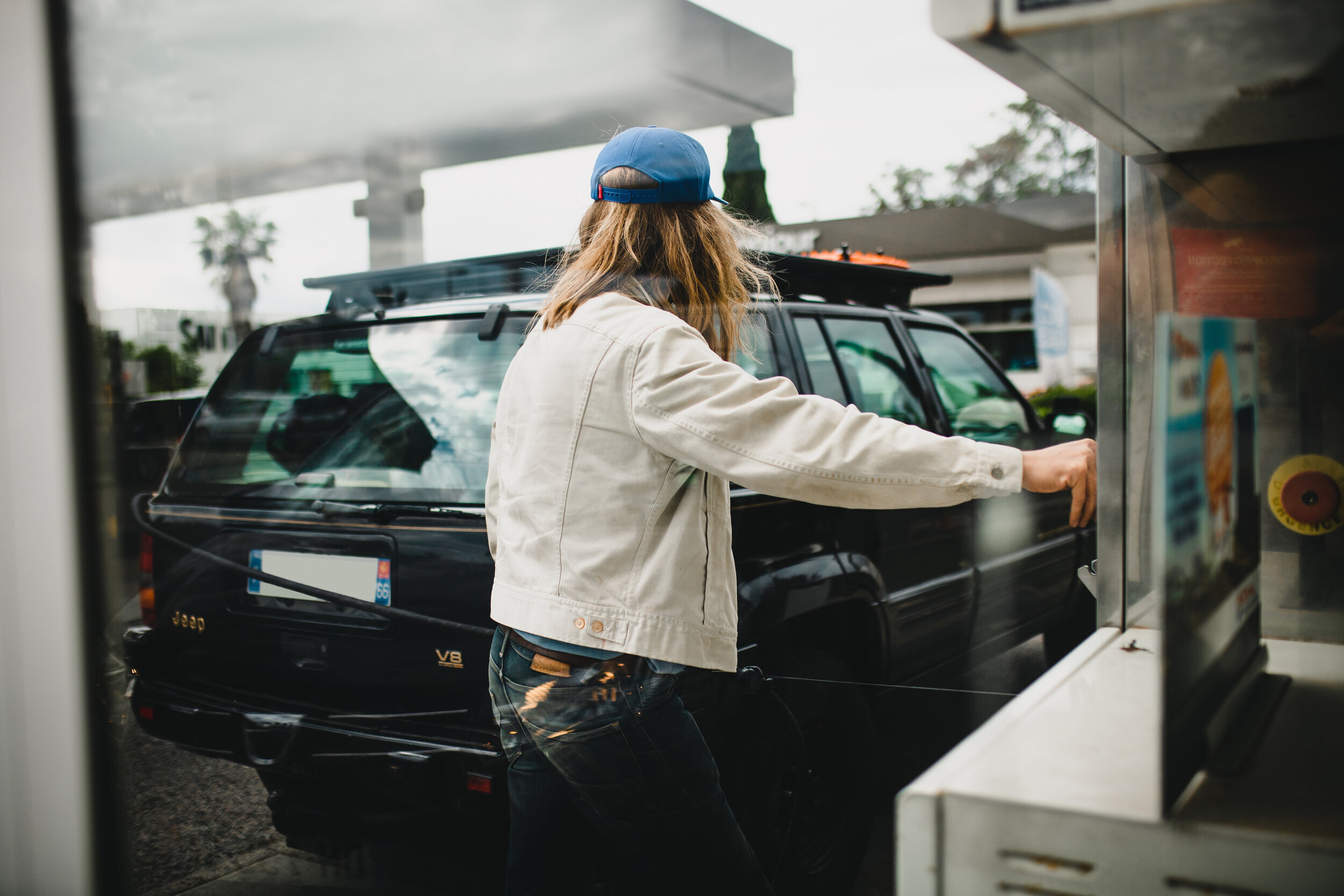 Personne rechargeant de l'essence dans une Jeep noire à une station-service, portant une veste blanche et une casquette bleue.