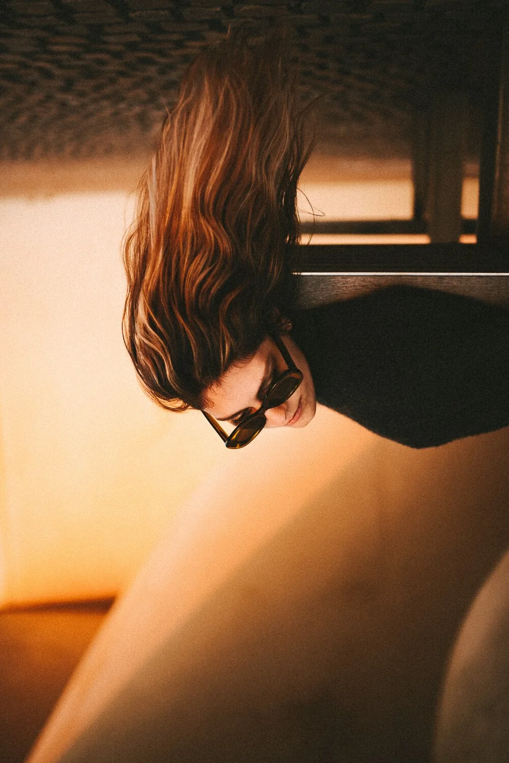 A woman with medium-length wavy brown hair and sunglasses is looking down, with her face partially visible. She is wearing a black top and is indoors, with warm lighting in the background.