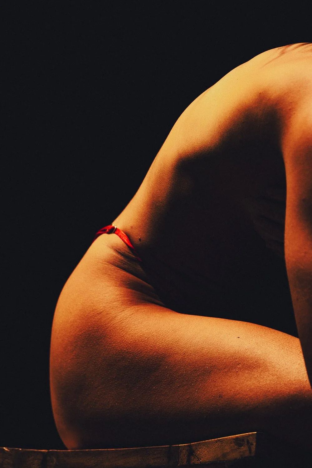 Close-up of a nude woman with fair skin in a contorted pose, with lighting highlighting her back, shoulder, and arm against a dark background.