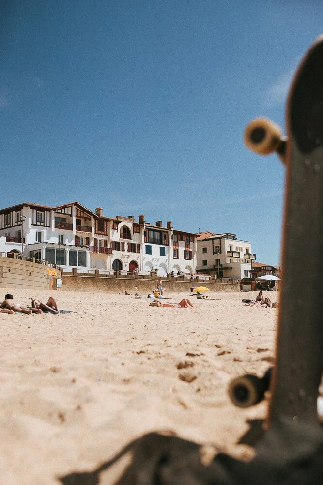 View of a sandy beach with people sunbathing and relaxing under umbrellas, overlooking multi-story beachfront houses against a blue sky, with part of a skateboard visible in the foreground.