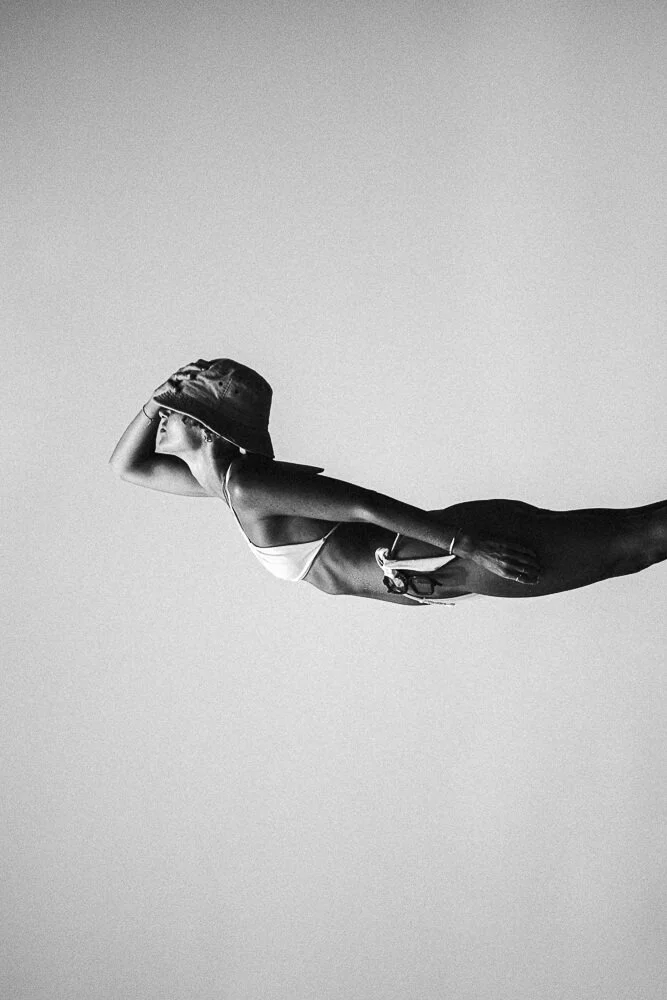 A black-and-white photo of a woman in a swimsuit lying horizontally, wearing a hat, with her hand shielding her eyes.