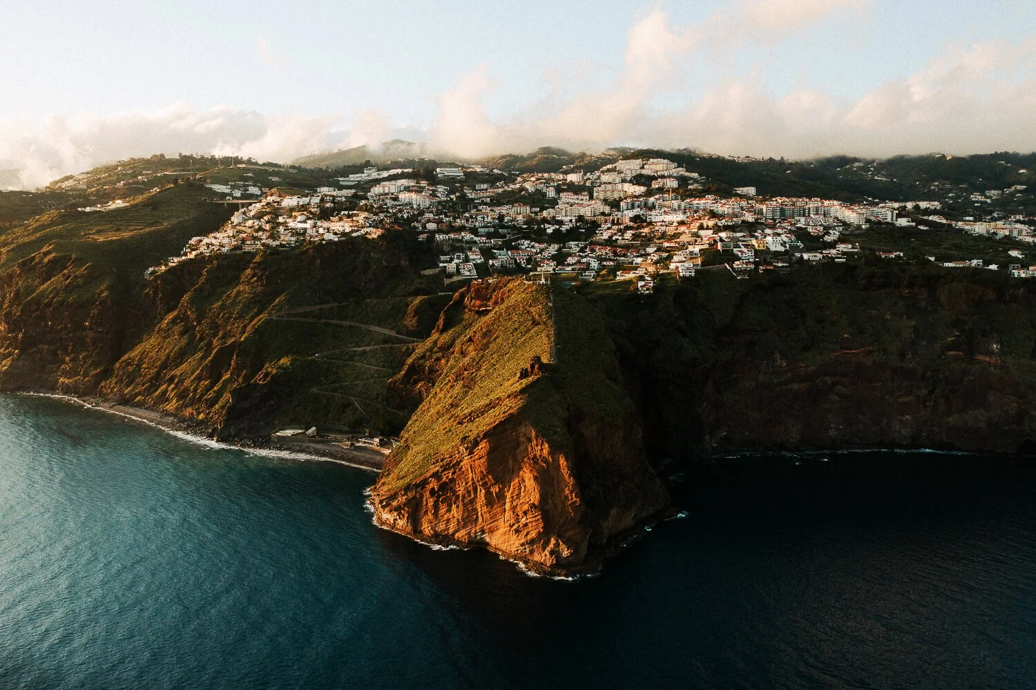 Aerial view of a coastal hillside town with white buildings, green vegetation, and a large rocky promontory extending into the ocean at sunset.