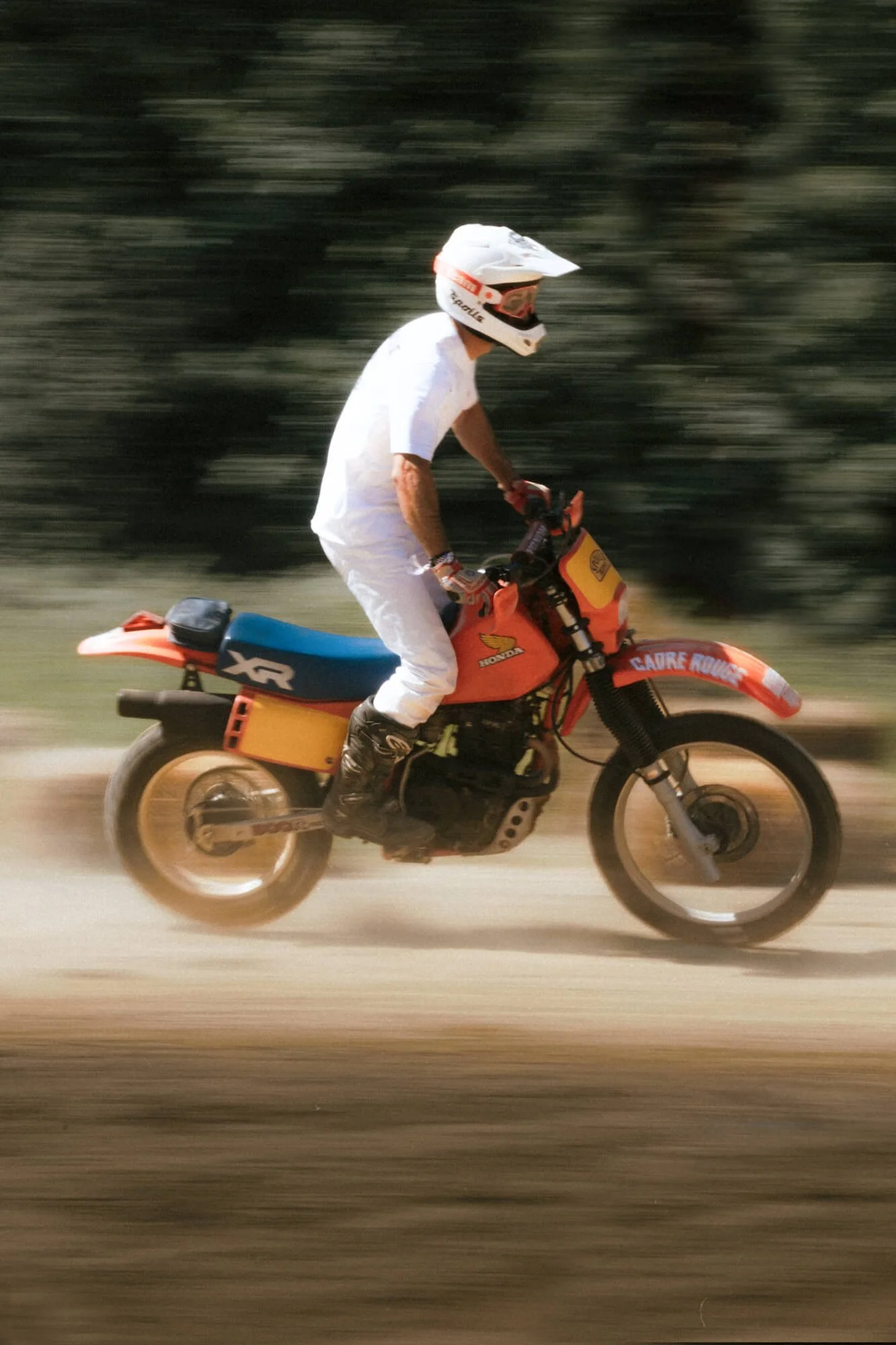 A person in white riding a red and yellow Honda dirt bike on a dirt trail, wearing a white helmet and gear, with a blurred background suggesting motion.