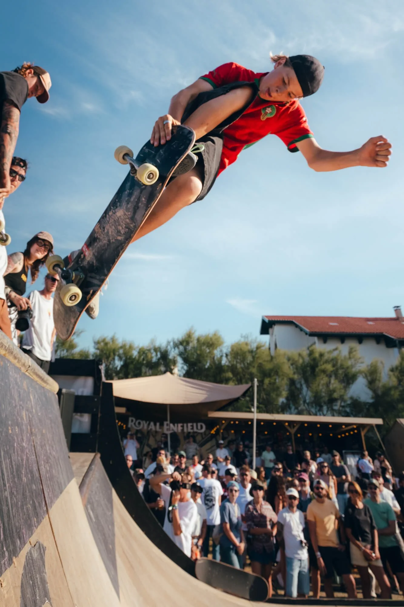 Skateboarder doing a trick on a ramp at an outdoor event, with a crowd watching. The sky is clear and sunny, and there are tents and buildings in the background.