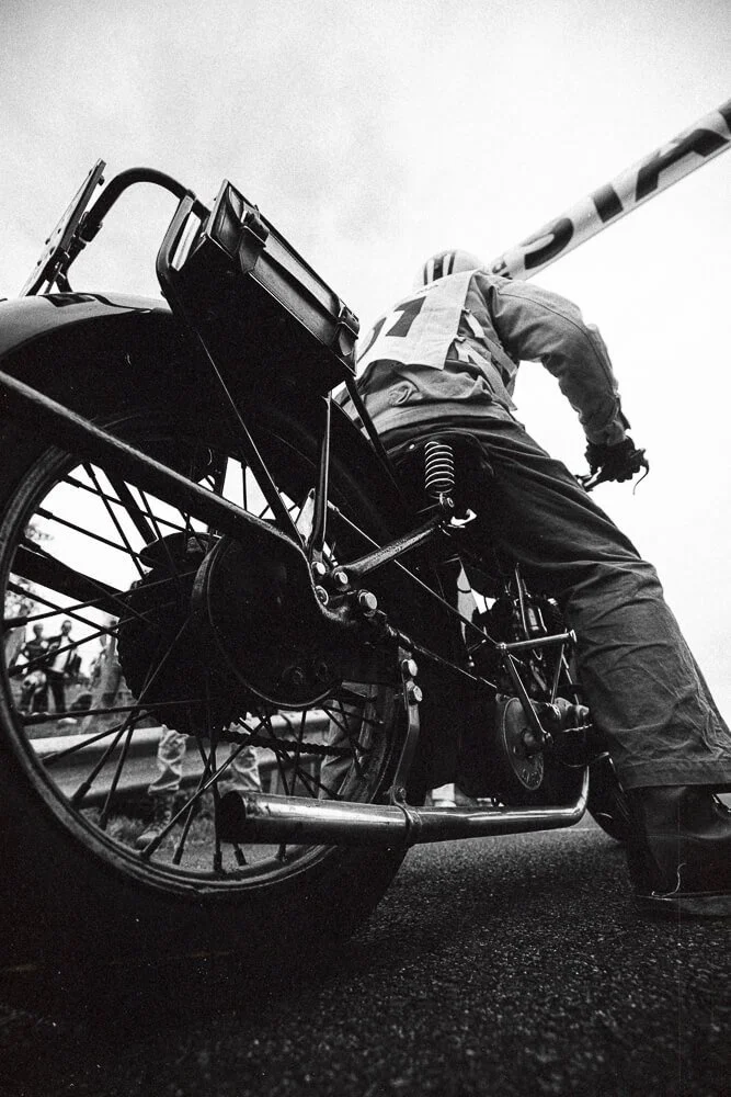 Close-up of a vintage motorcycle and a person leaning over it, possibly at a race or event, in black and white.