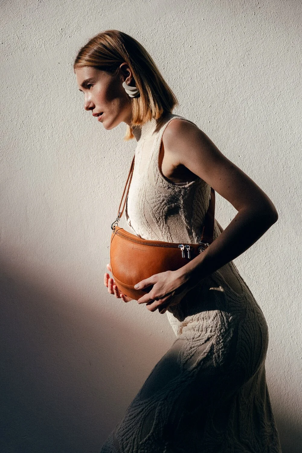 A woman with shoulder-length hair, wearing a sleeveless textured dress and white earrings, stands sideways against a plain wall while holding a brown leather bag in front of her waist.