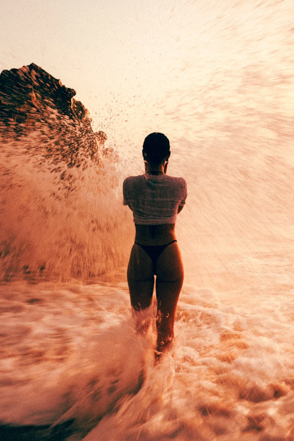 A woman in a bikini standing in the ocean with a breaking wave on one side and a rock formation in the background, during sunset.