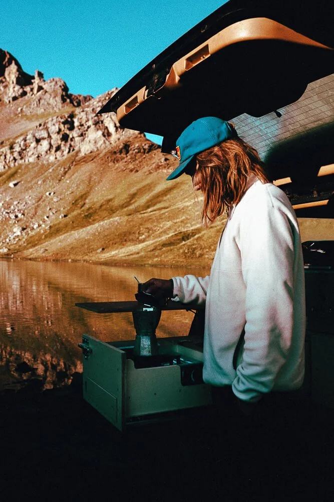 Person with orange hair wearing blue cap and white jacket operating a drone controller near a mountain lake.