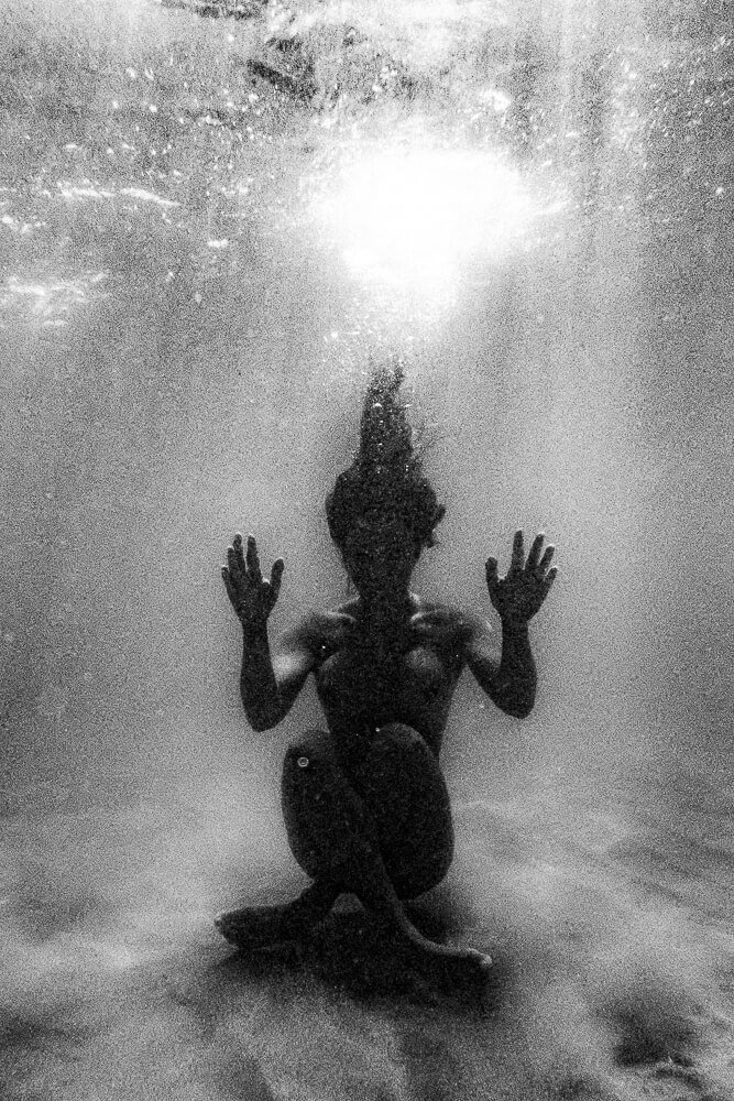 A person underwater, sitting cross-legged with arms raised, facing the camera, with sunlight filtering down from above.
