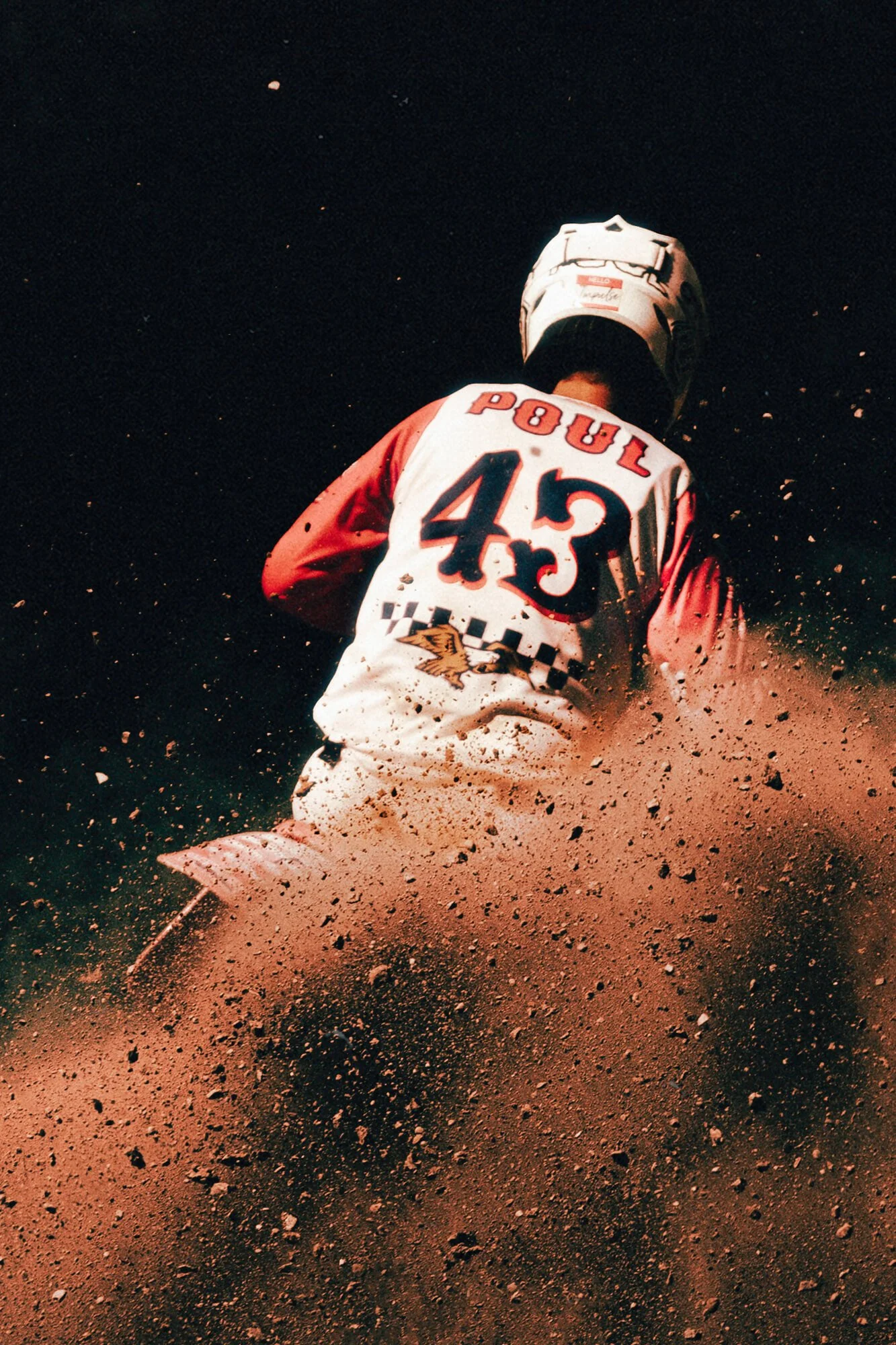 A dirt bike rider wearing a white jacket with P-81 and 43 on the back, and a white helmet, riding on a dirt track kicking up dust and small rocks.