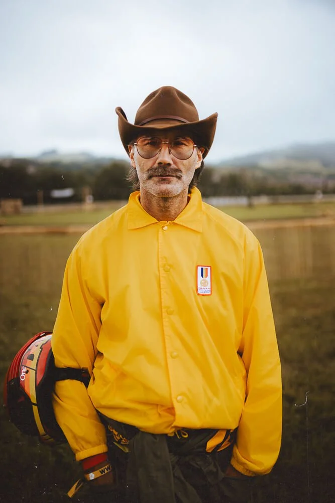 A man wearing a brown cowboy hat, sunglasses, and a yellow jacket standing outdoors on a cloudy day.