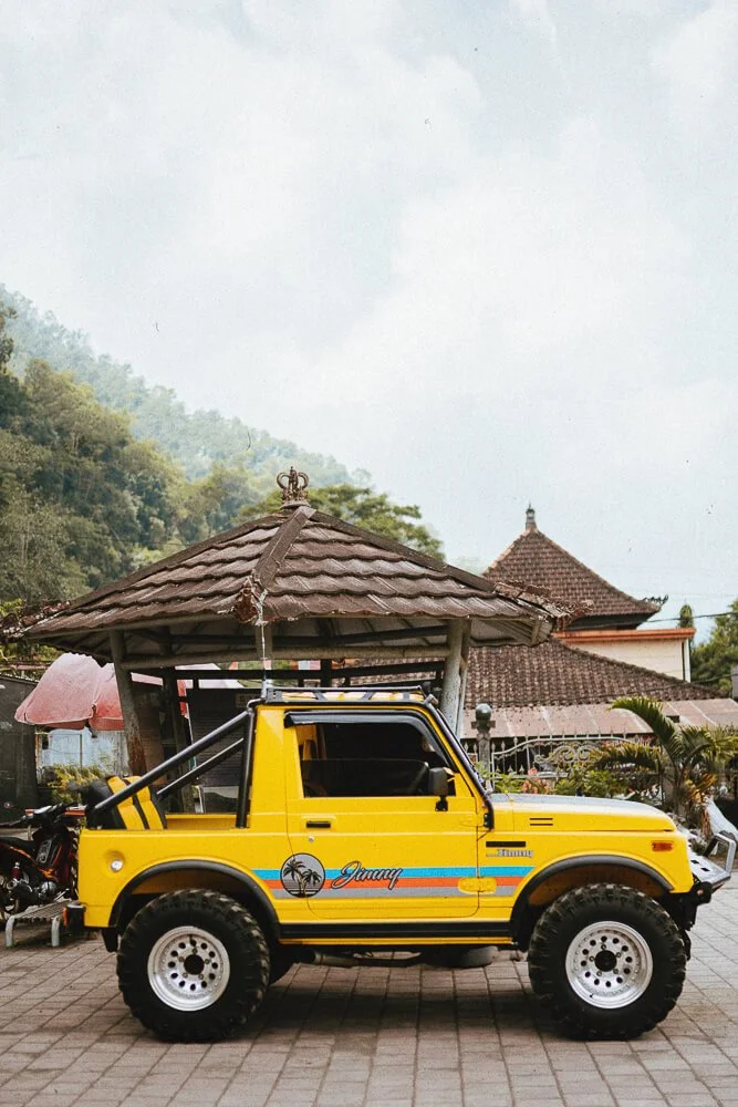 Yellow off-road vehicle with painted palm trees and the name "Jimmy" parked on a paved area in front of traditional buildings with tiled roofs and lush green trees.