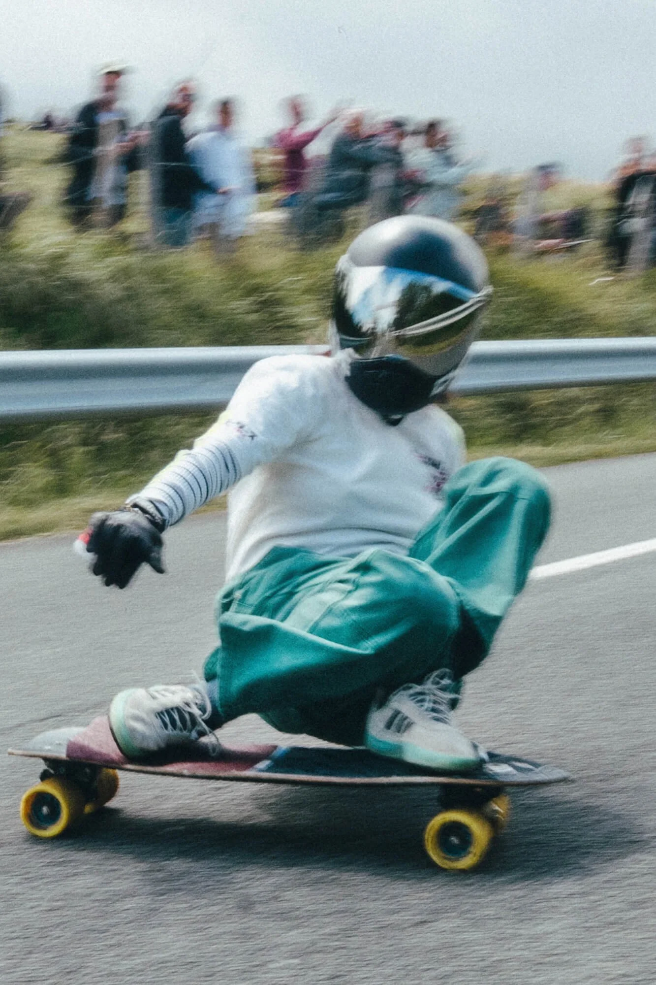 A person wearing a white shirt, teal pants, and a black helmet is skateboarding on a paved road. The skateboard has yellow wheels. The person is leaning forward, and there is a group of spectators blurred in the background.