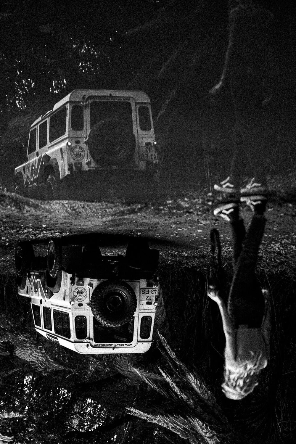 Black and white photo of a Land Rover parked on a dirt trail among trees, with its reflection visible in a puddle in the foreground. A person wearing athletic shoes is standing nearby.