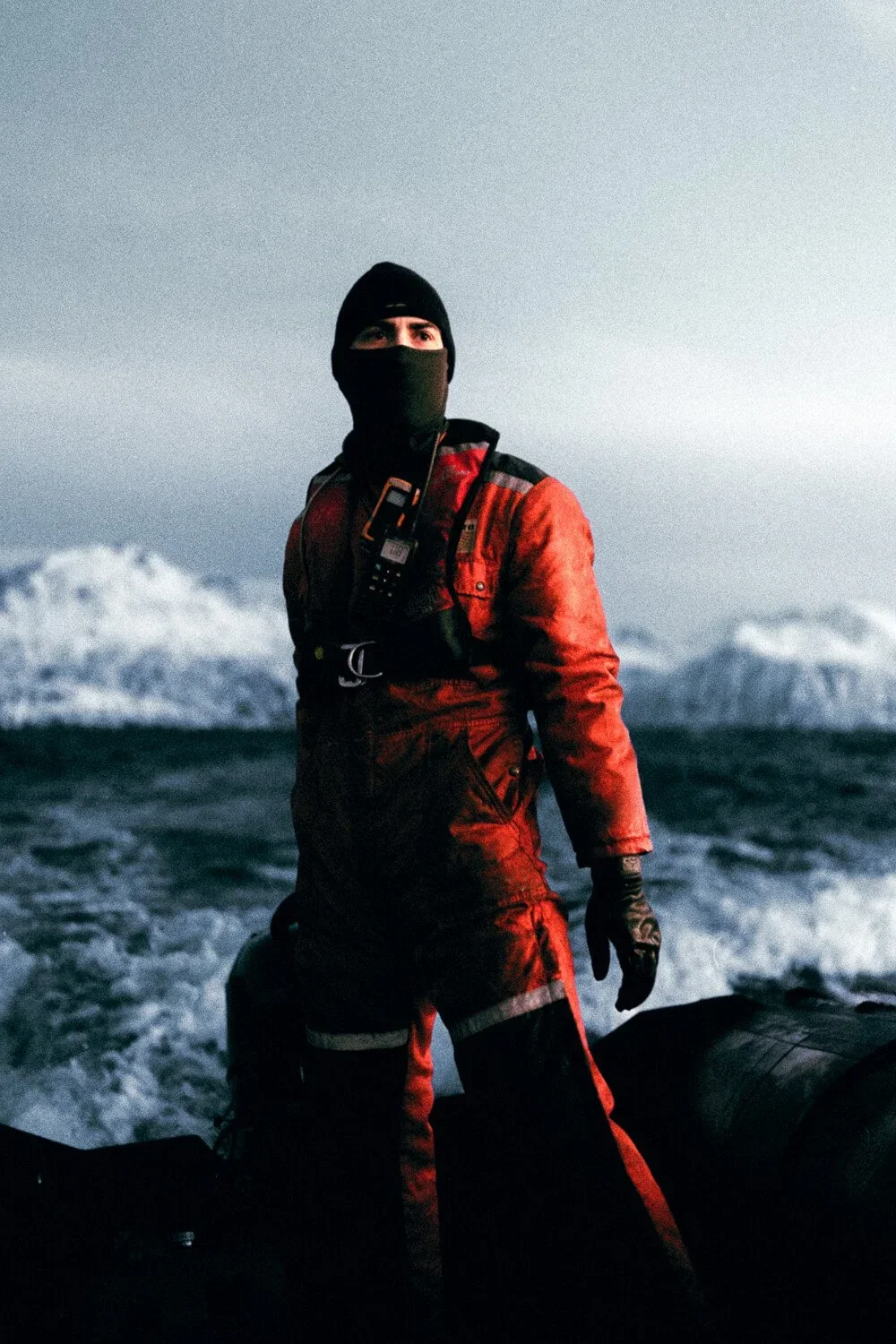 Un homme en tenue de sauvetage en mer, portant un casque et un masque faciale, se tenant sur un bateau de sauvetage dans une mer agitée avec des montagnes enneigées en arrière-plan.