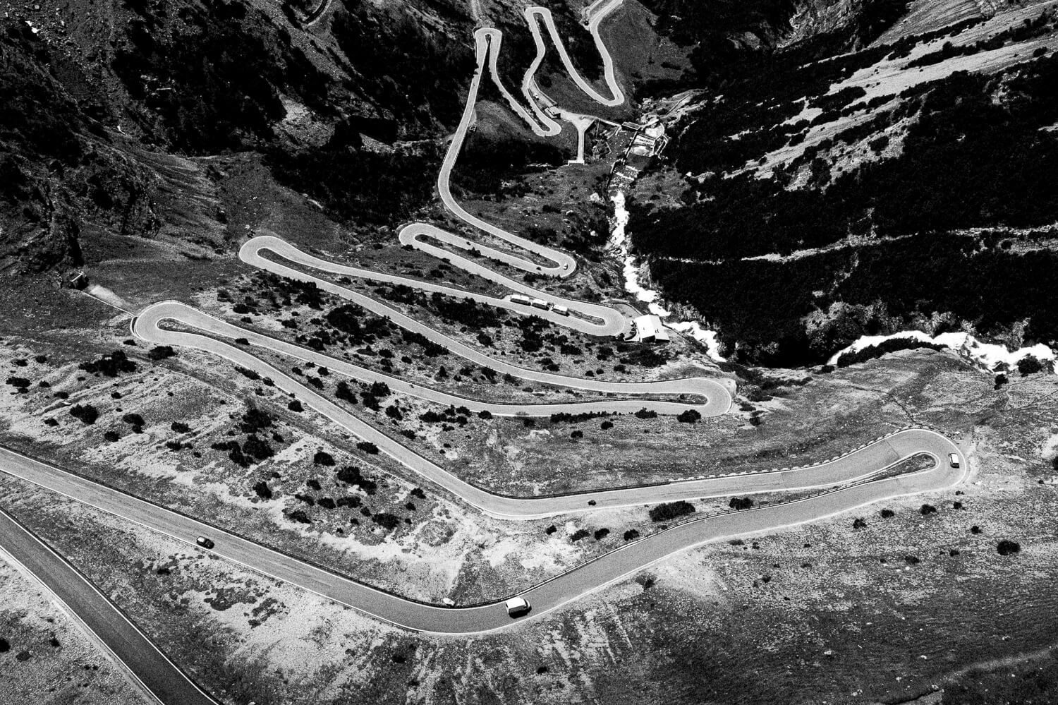 Black and white aerial photograph of a winding mountain road with multiple switchbacks. Several vehicles are driving along the road, and a river runs through the valley below.