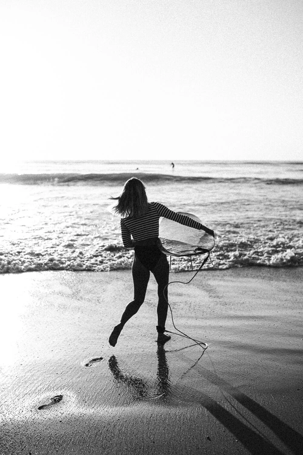 A woman on the beach holding a surfboard, facing the ocean during sunset, in black and white.