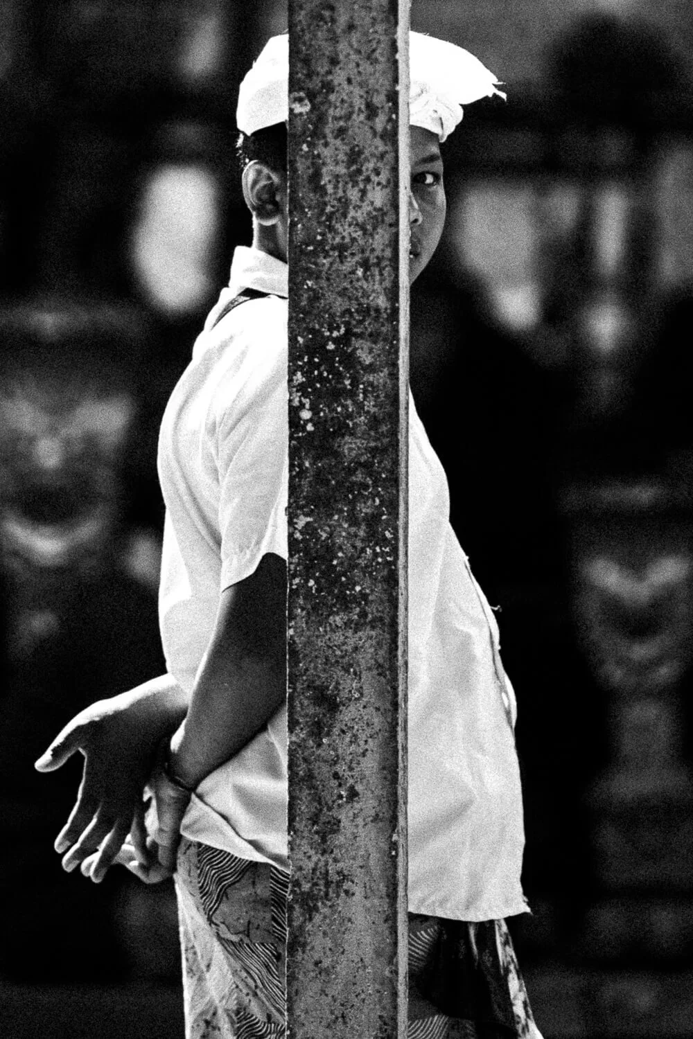 Une jeune fille avec un vêtement blanc, portant un bandeau blanc sur la tête, se tient derrière un poteau en fer rouillé, regardant vers l'appareil photo. La photo est en noir et blanc.