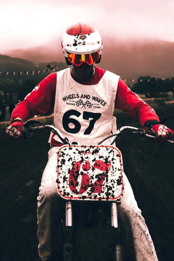 Motocross rider wearing a white helmet with red accents, red goggles, and red and white racing gear, sitting on a dirt bike with a number 61 on the front plate, covered in mud with a blurred outdoor landscape in the background.