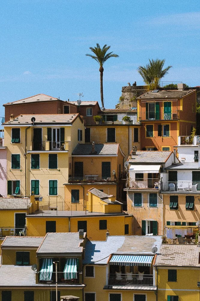 Colorful Mediterranean apartment buildings with green shutters, balconies, and tiled roofs, set against a bright blue sky with two tall palm trees in the background.