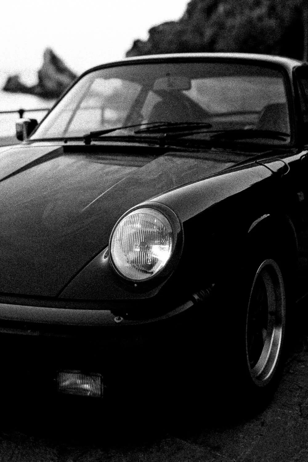 Black and white photo of a classic sports car with round headlights, parked near a coastal area with rocky cliffs in the background.
