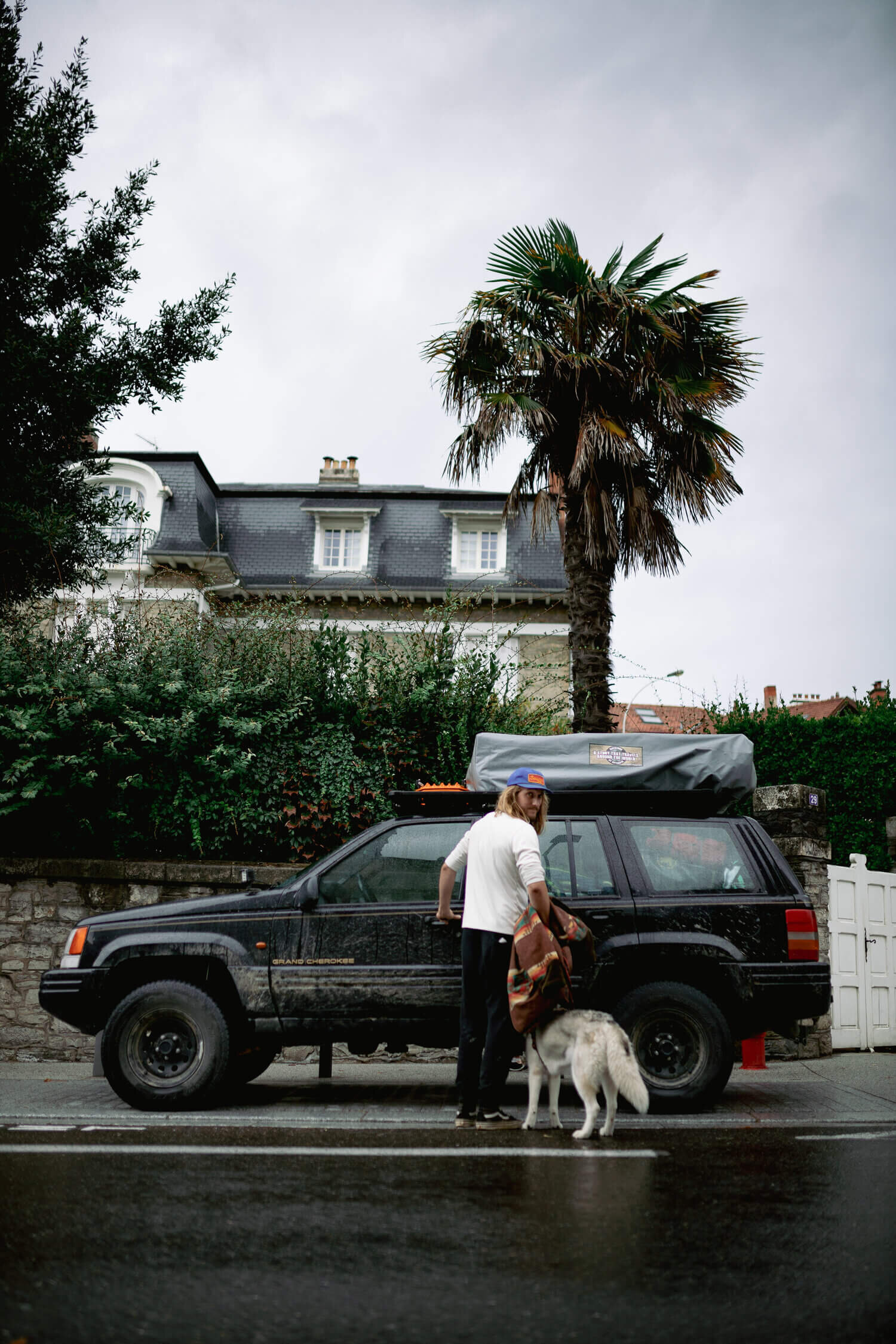 Un homme portant une casquette bleue se tient à côté d'un SUV noir avec une tente de toit. Un chien blanc et gris se trouve à ses pieds. Derrière, il y a un palmier, des buissons verts et une grande maison de style traditionnel. Le sol est mouillé, s
