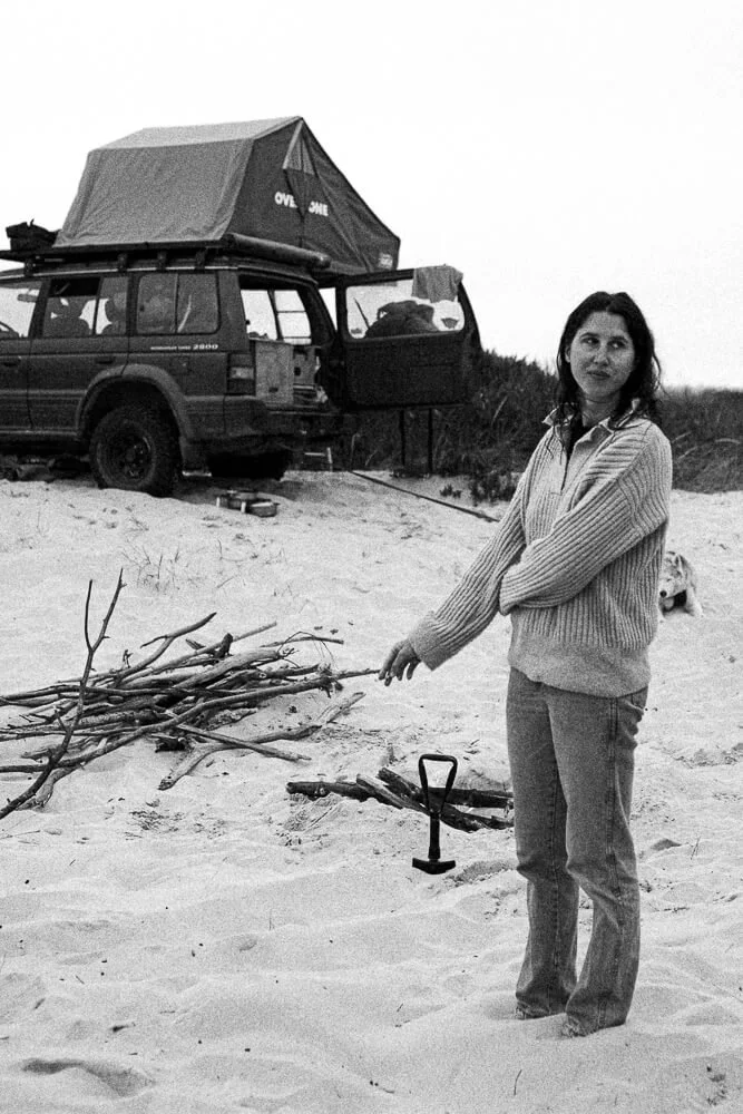 A woman standing on a sandy beach holding a small shovel, with a pile of driftwood nearby, and a vehicle with a rooftop tent in the background.