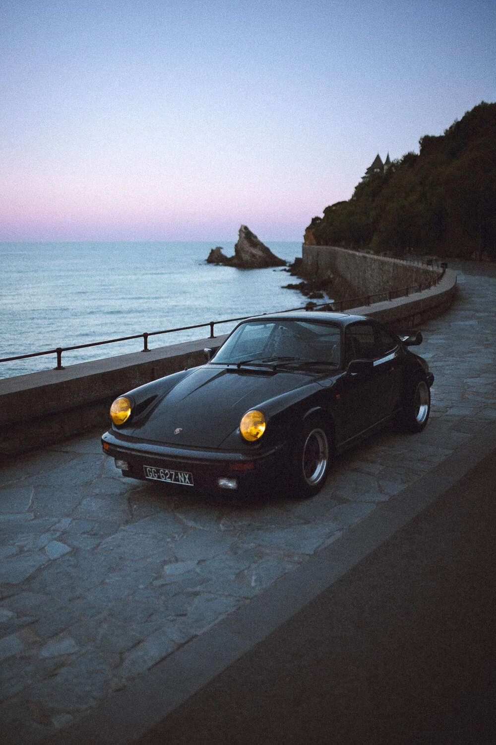 A black vintage Porsche 911 parked on a stone-paved coastal road at dusk, with the sea, rocks, and a rocky coastline under a pink and blue sky in the background.