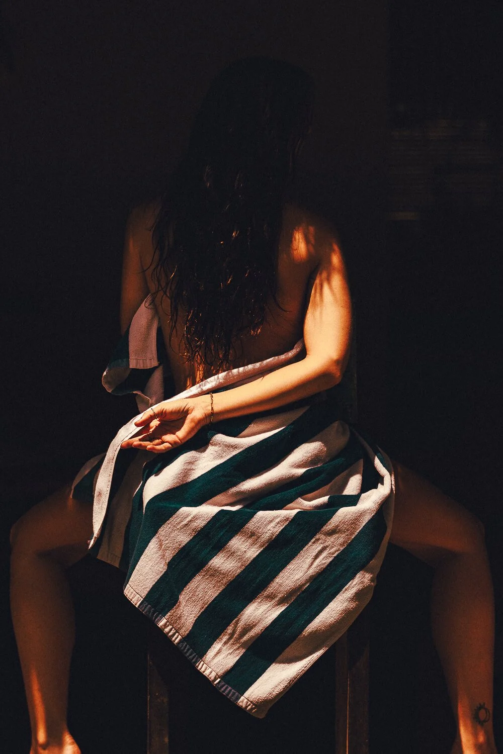 A person sitting on a chair with their legs apart, wrapping a towel around their waist, with long dark hair covering their face, in a dimly lit environment.