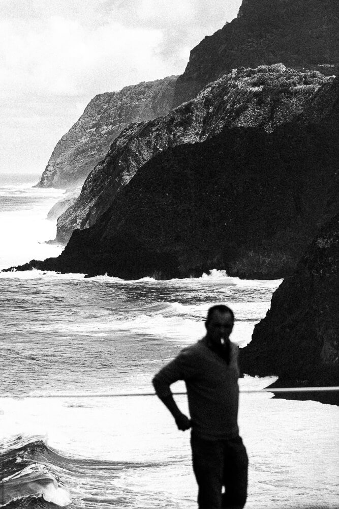 A blurry man standing on a beach with dramatic cliffs and ocean waves in the background in black and white.