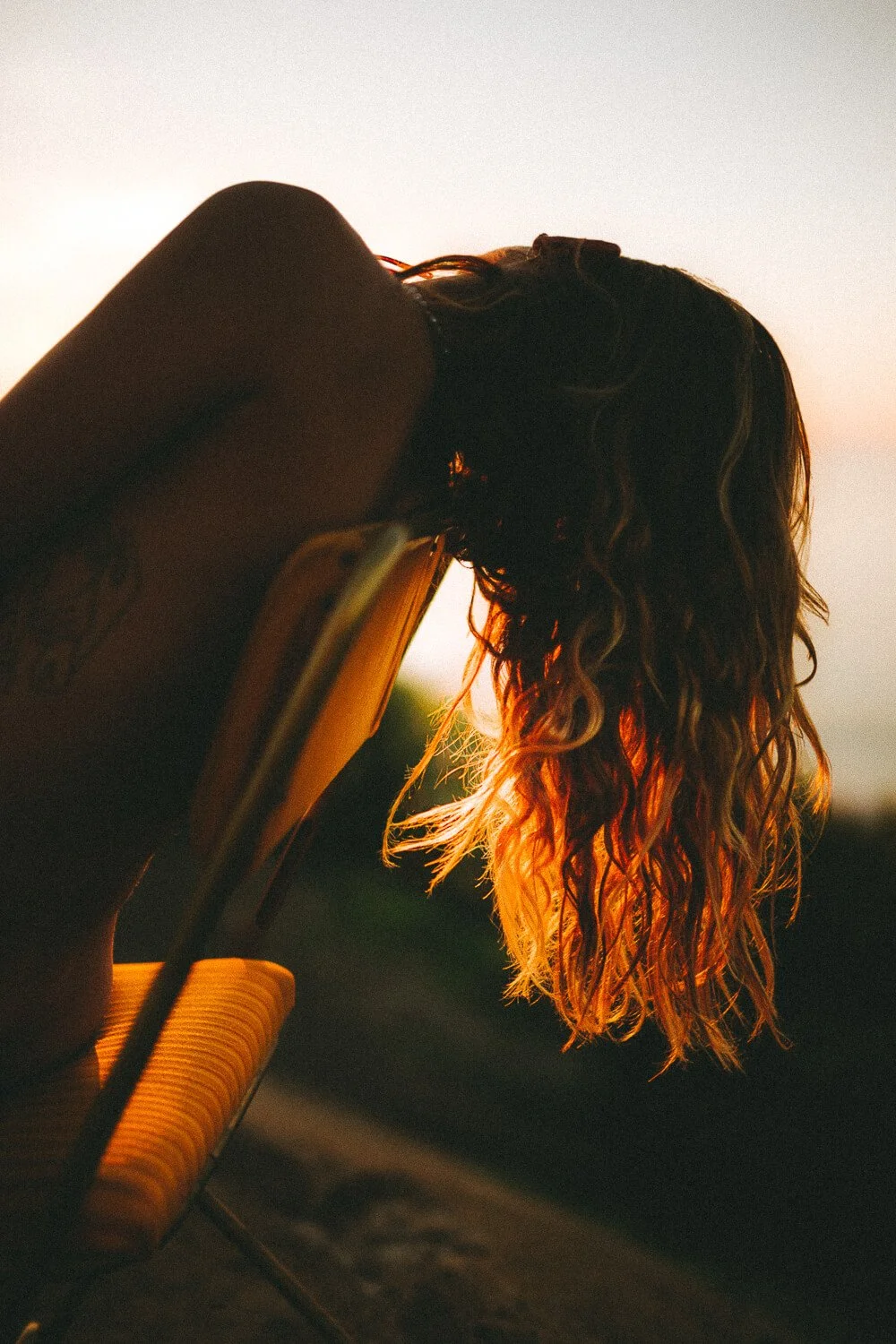 Person with wavy hair seated on a chair, backlit by sunset.