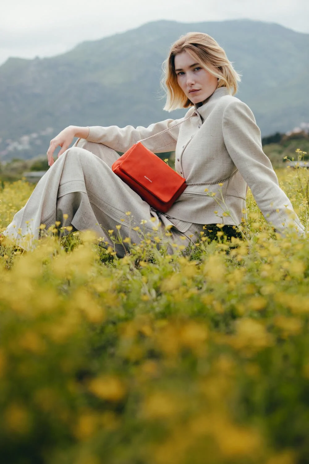 A woman sitting in a field of yellow flowers outdoors, wearing a beige outfit, with a red purse hanging from her shoulder, and mountains in the background.
