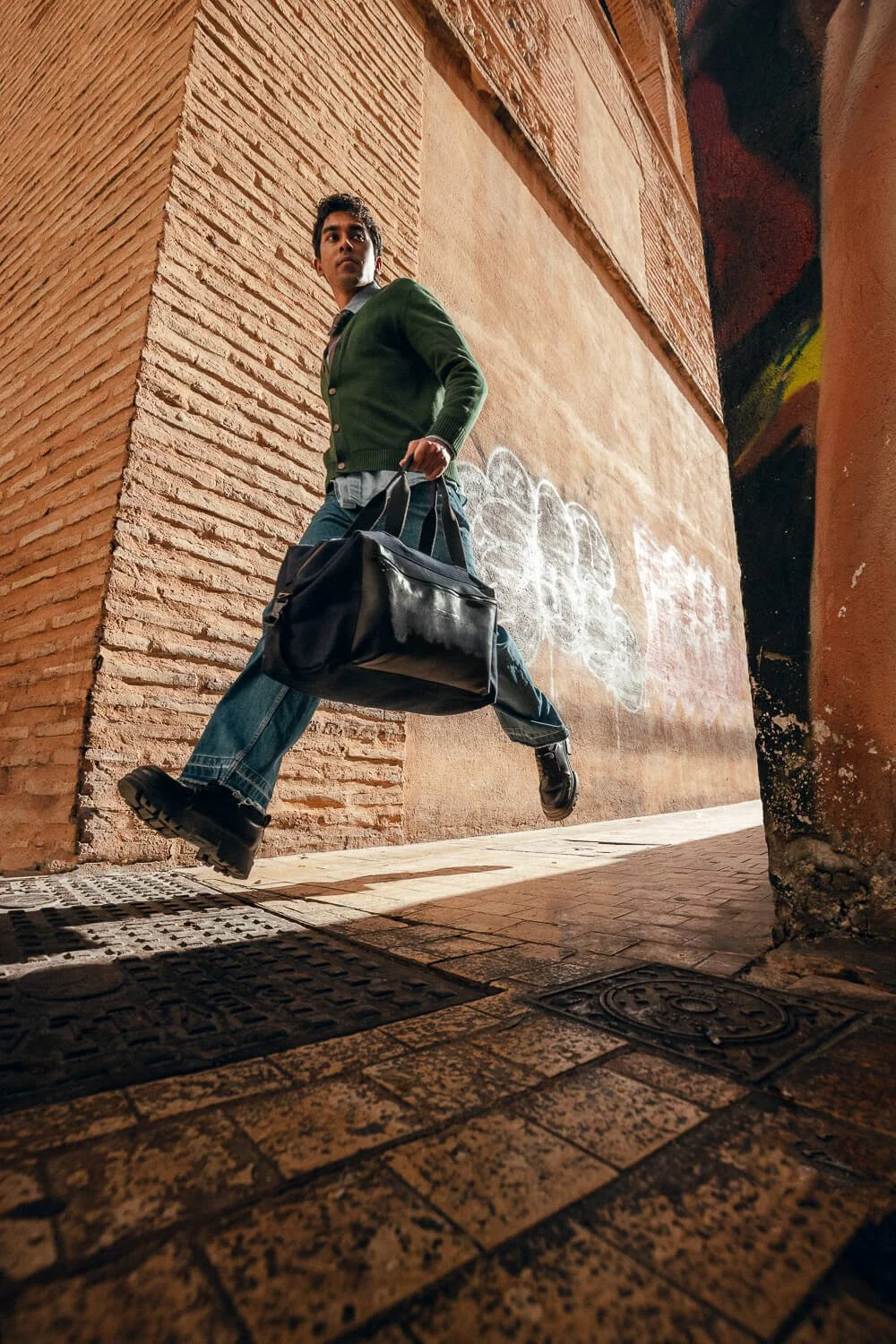 Young man walking quickly through a narrow alleyway, carrying a black duffel bag, with brick and graffiti-covered walls.