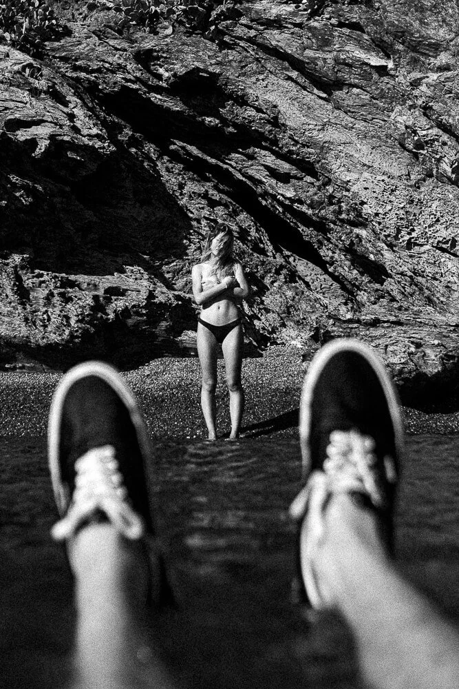 A person in sneakers is lying in water at the beach, taking a photo of a woman in a black bikini standing near rocks.