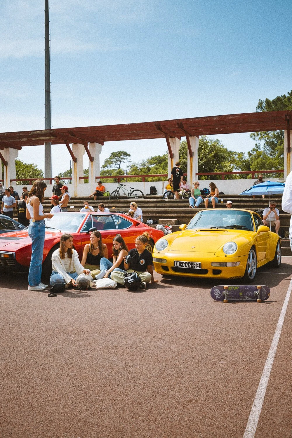 Group of young people sitting on the ground near a yellow Porsche and a red sports car at an outdoor car meet on a sunny day.
