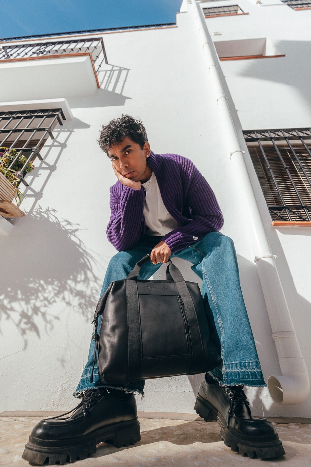 A young man with curly hair sitting outside against a white building, dressed in a purple cardigan, white shirt, blue jeans, and black boots, holding a black bag and resting his face on his hand.
