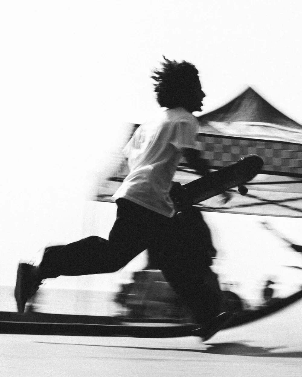Une personne en mouvement, tenant une planche de skateboard, en noir et blanc.