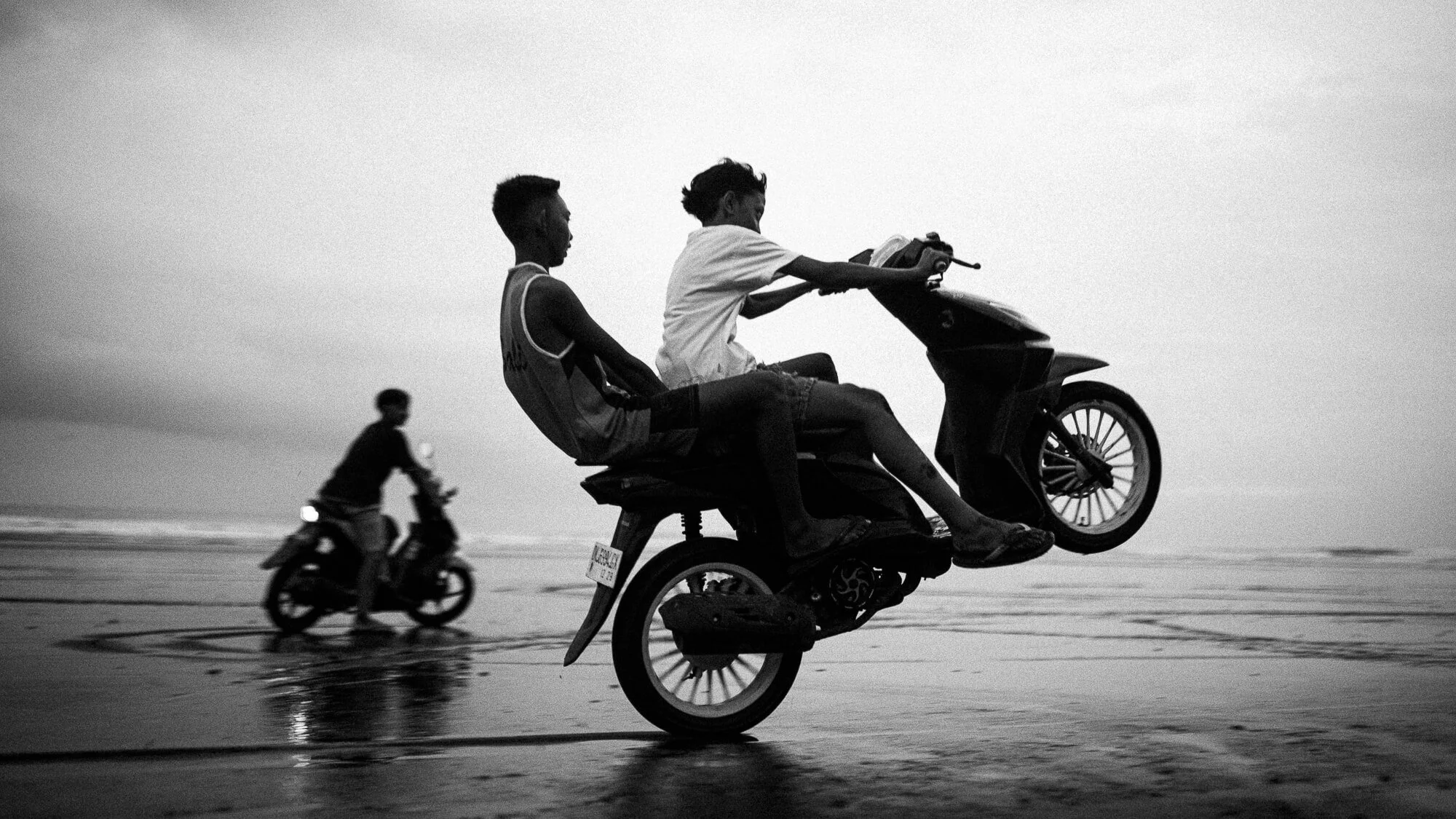 Deux jeunes garçons sur une moto, l'un en train de faire un wheeling sur la plage, avec un autre garçon en moto derrière eux. Image en noir et blanc.