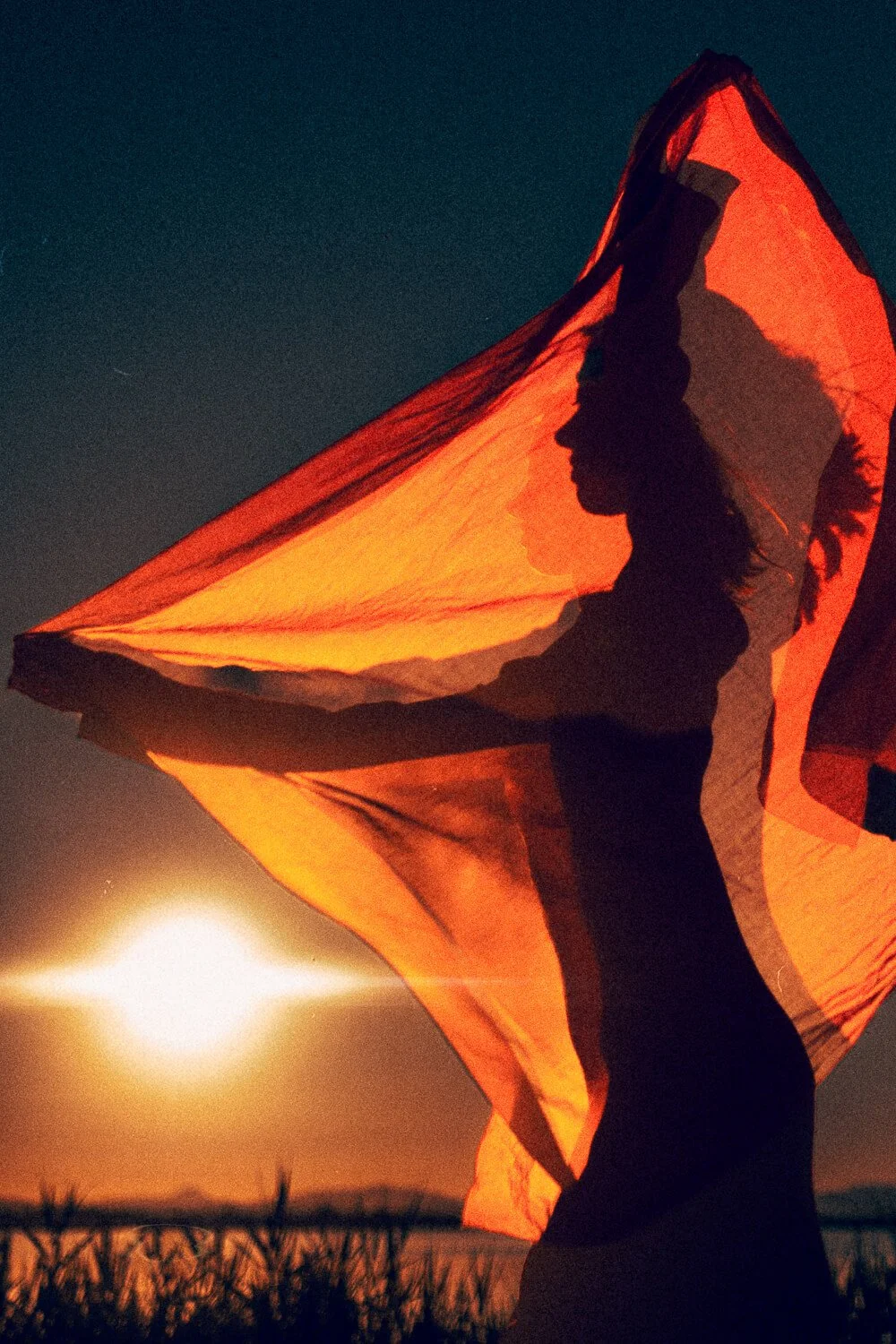 A woman holding a large orange fabric, silhouetted against a sunset sky with the moon visible in the background.