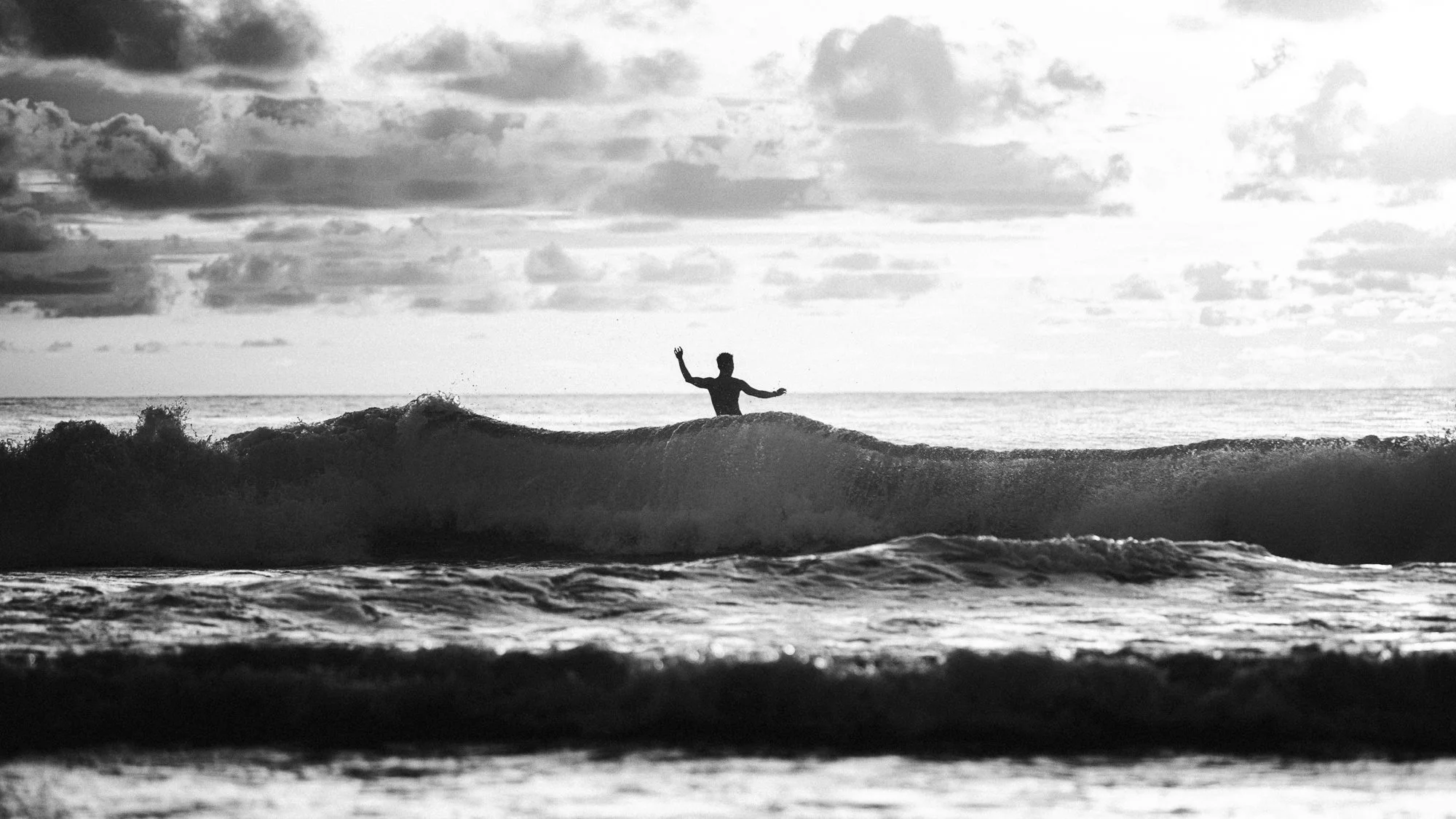 Un surfeur en silhouette se tient debout sur une vague à la plage, sous un ciel nuageux.