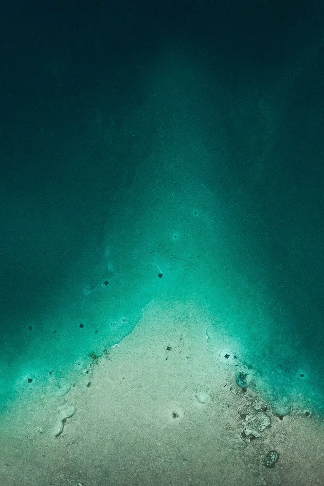 Underwater view of a sandy ocean floor with scattered small rocks and debris, where the water appears clear and teal.