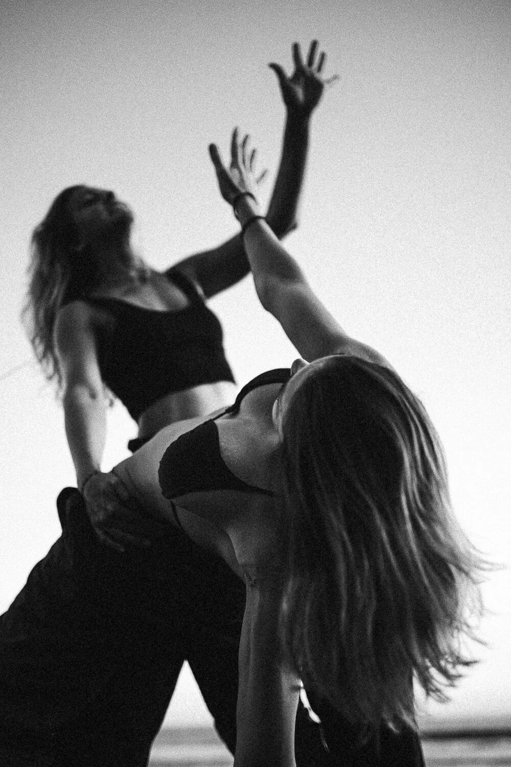 Two women are dancing close to each other with one reaching up and the other leaning back, on a beach at sunset or sunrise, in black and white.