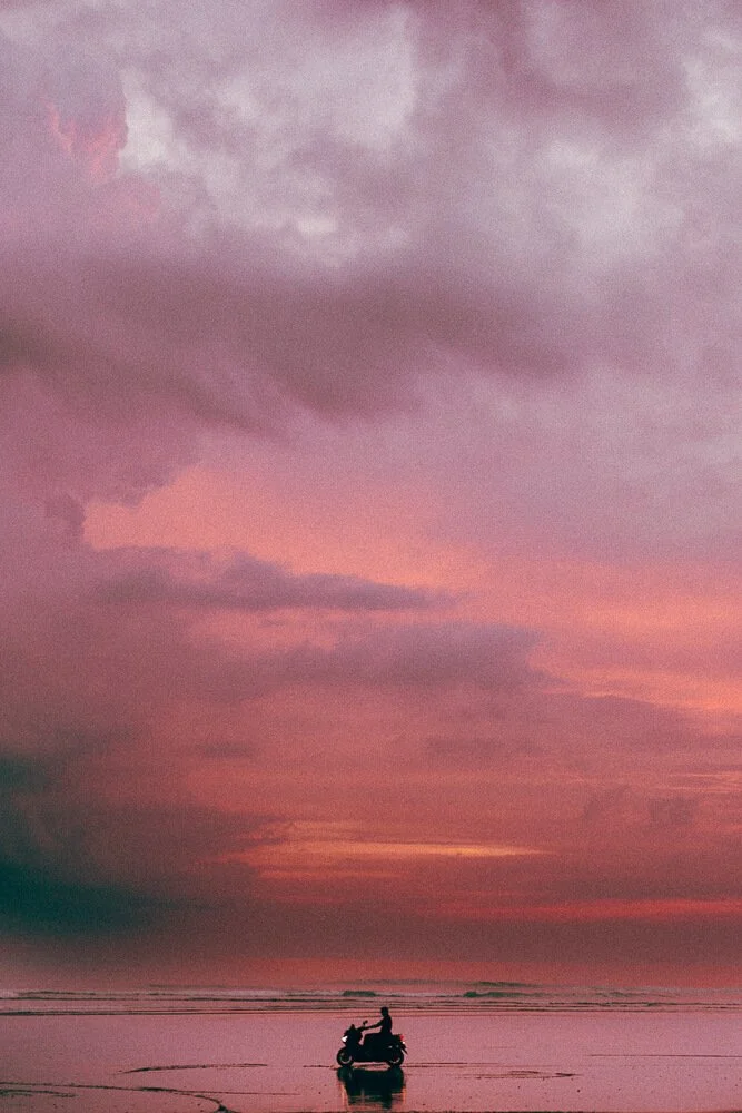 Person riding a scooter on the beach at sunset with a colorful purple and pink sky.